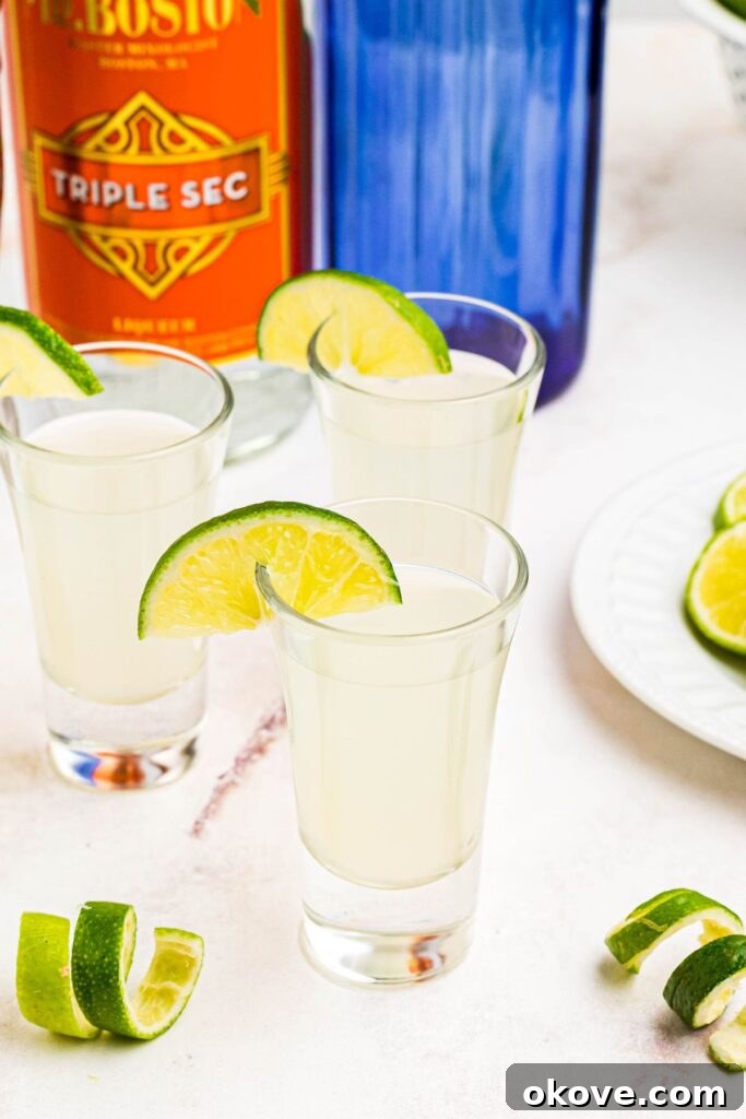 Crisp and refreshing Kamikaze shots in elegant shot glasses, garnished with vibrant lime wedges and artfully curled lime peels, all presented on a luxurious marble table, ready for a sophisticated gathering.