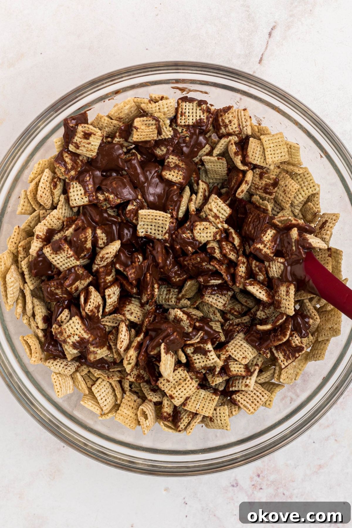 Chocolate and peanut butter mixture being mixed with Chex cereal in a large clear glass bowl. 