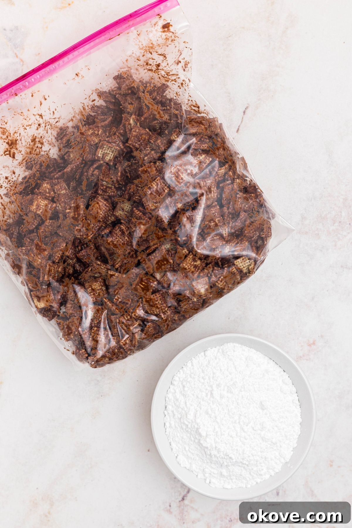 Chocolate coated cereal in a large bag, with a bowl of powdered sugar next to the bag, before mixing together. 