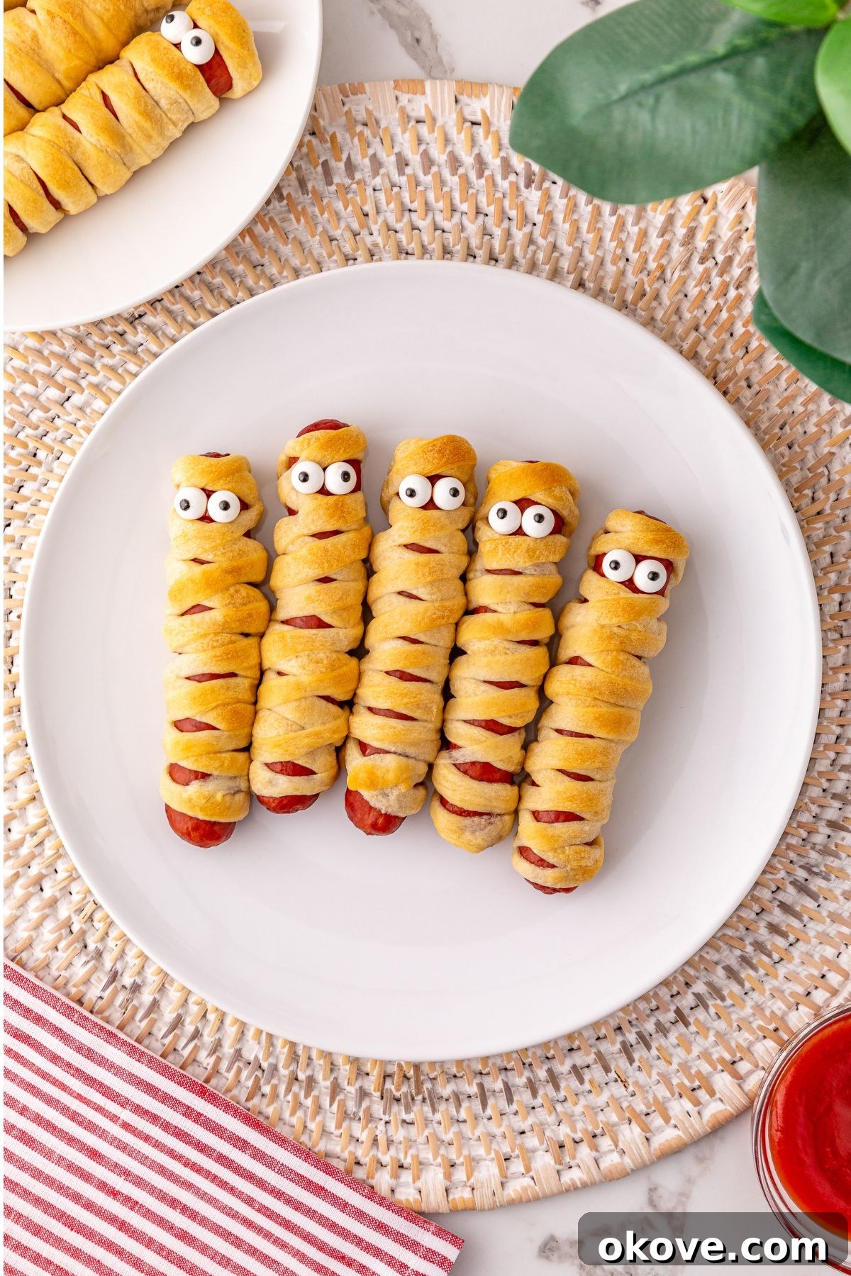 Mummy Hot Dogs 2 Mummy hot dogs artfully arranged on a pristine white plate, adorned with whimsical candy eyes, ready for a Halloween celebration.