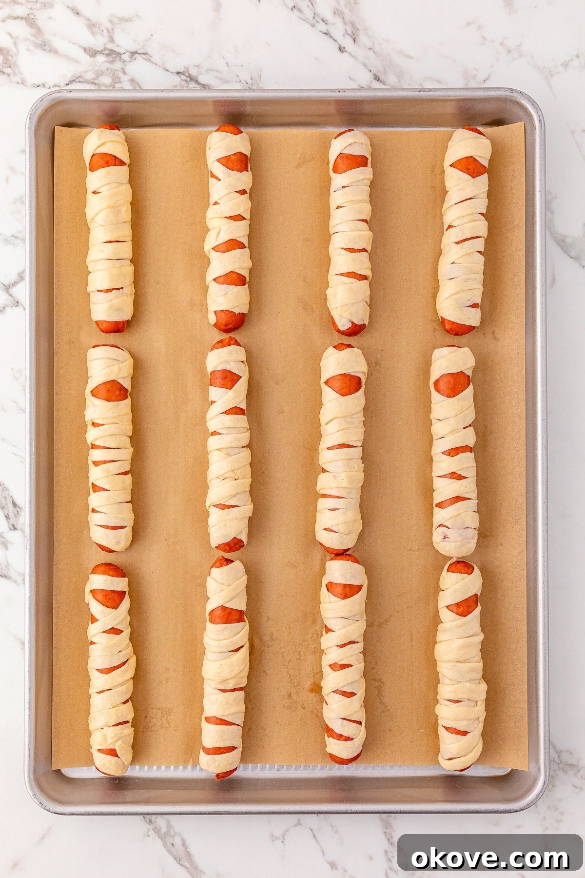 Mummy Hot Dogs 5 Uncooked hot dogs carefully wrapped with strips of crescent dough, arranged neatly on a baking sheet lined with parchment paper, awaiting baking.