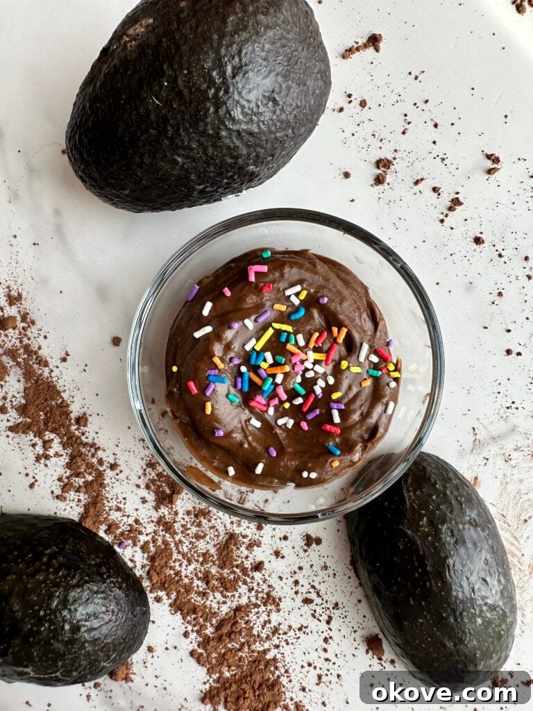 A glass bowl filled with creamy avocado chocolate pudding, adorned with colorful sprinkles, sits on a rustic wooden table. Fresh, halved avocados are artfully placed around the bowl, emphasizing the natural, wholesome ingredients used in the dessert, perfect for healthy snacking.
