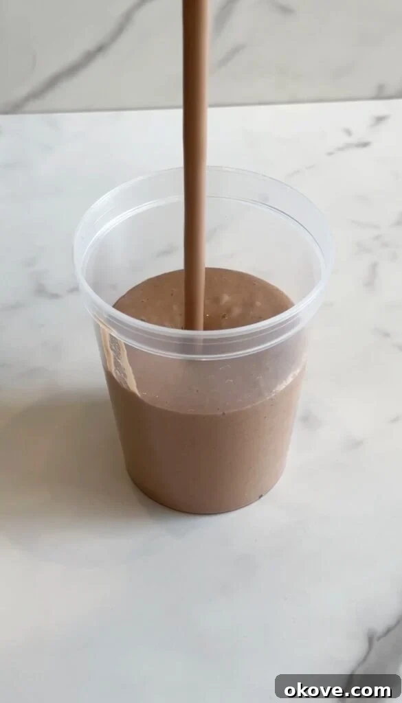The smooth and creamy chocolate spinach milkshake is being poured from a blender into a clear plastic cup, ready to be enjoyed.