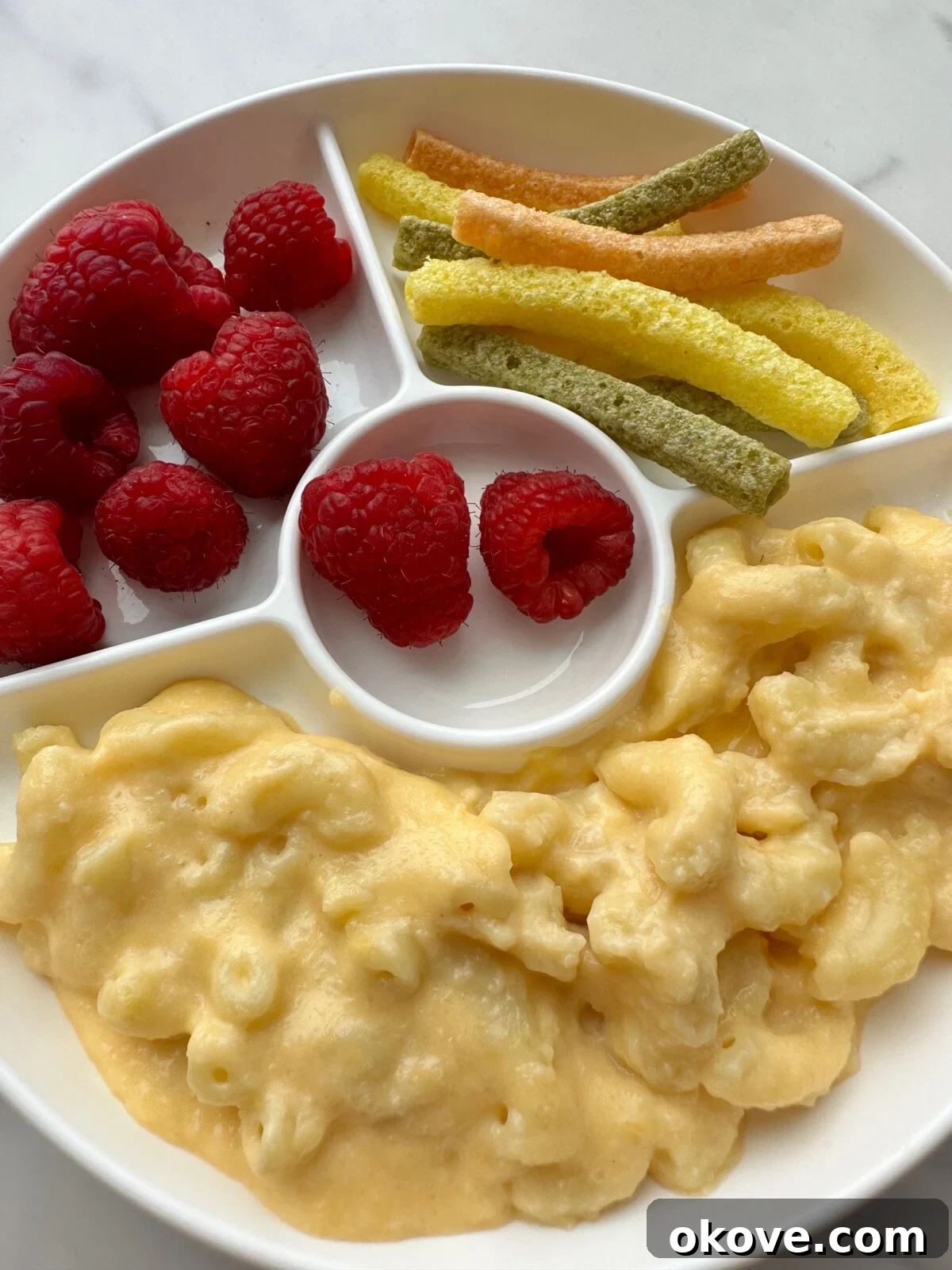 Irresistible Cauliflower Mac and Cheese 3 A close-up of the delicious Cauliflower Mac n Cheese in a divided plate, served with fresh raspberries and crunchy vegetable straws, a perfect balanced meal for children.