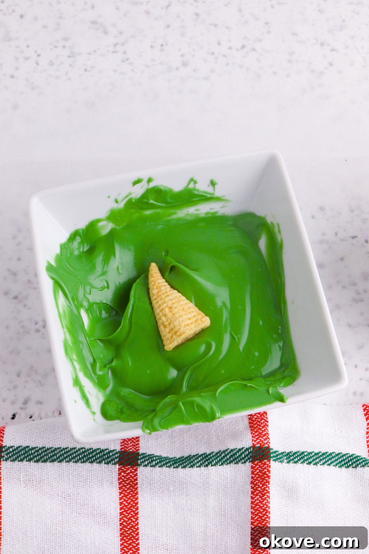 Elven Fanfare Headwear 4 A corn chip Bugle being carefully dipped into a square dish filled with smooth, melted green candy melts, ensuring a complete and even coating.