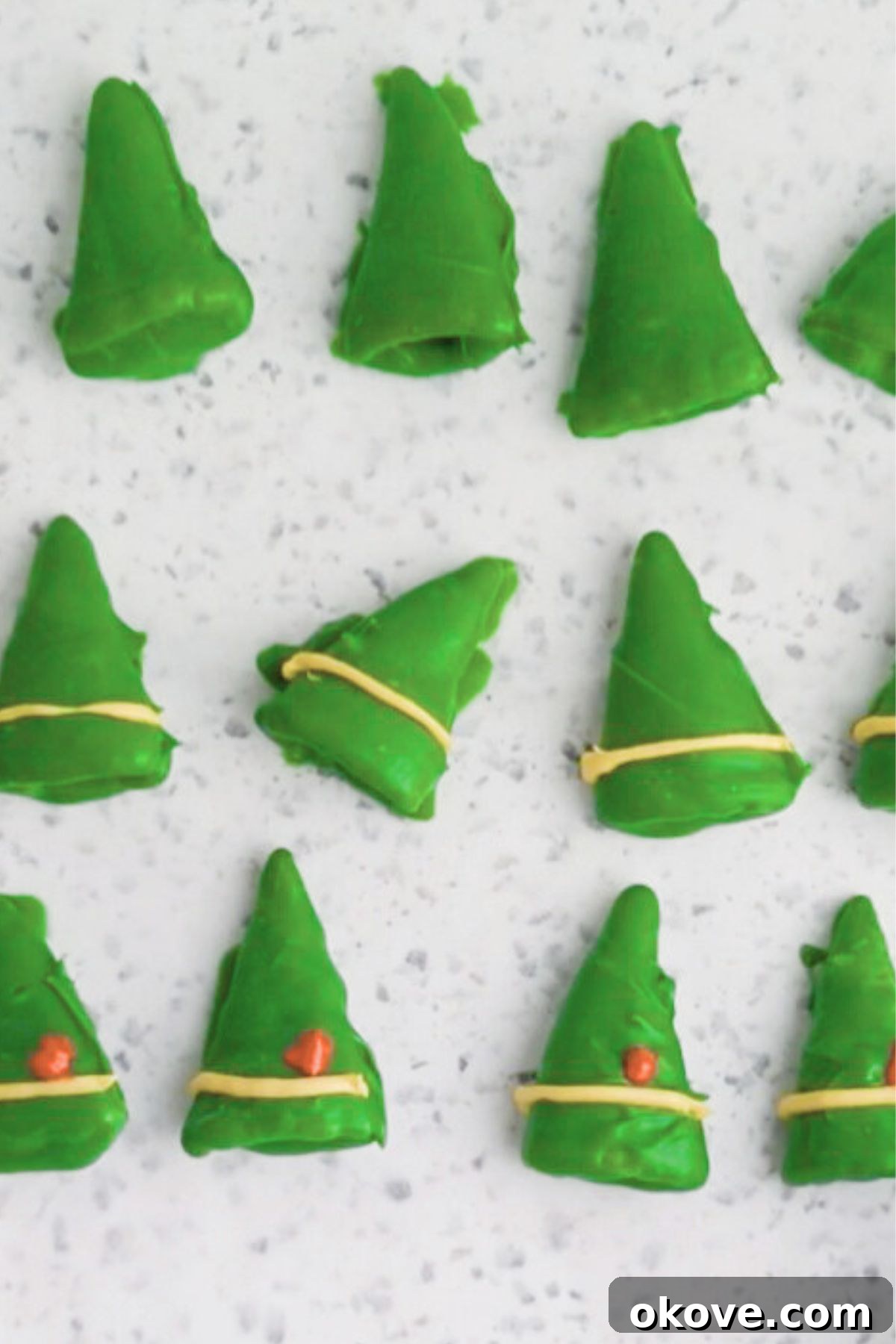 Elven Fanfare Headwear 5 Green coated Bugles, now dried on parchment paper, are being meticulously decorated with a thin stripe of yellow candy melts along the base and a small dot of red melts, transforming them into charming elf hats.