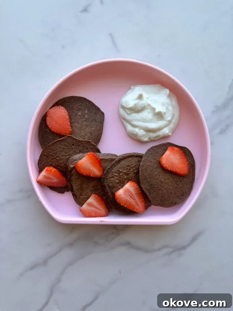 Hidden Greens Chocolate Pancakes 2 Five chocolate spinach pancakes on a pink plate with a side of whipped cream and topped with strawberries.