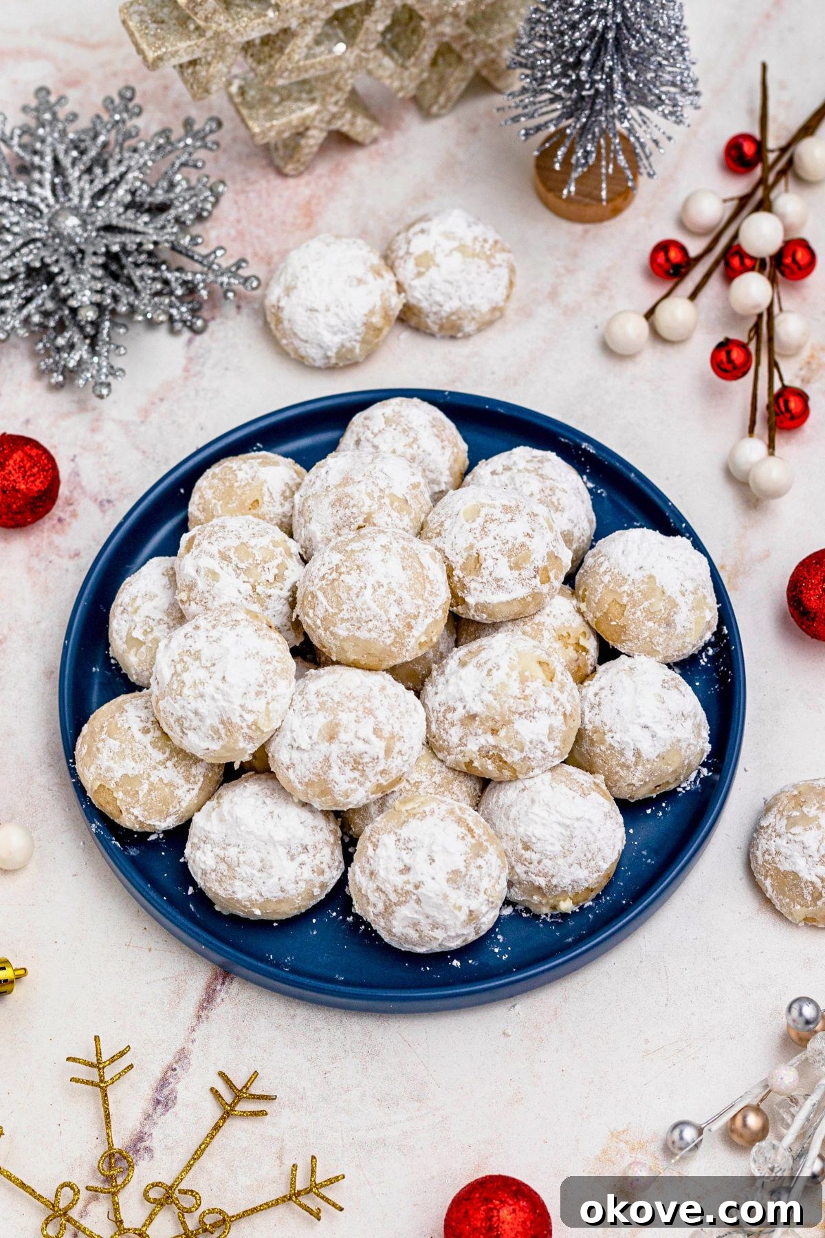 Powdered Cloud Kisses 2 Powder sugar coated Snowball cookies, stacked on a blue plate with holiday party favors on the table around the plate.