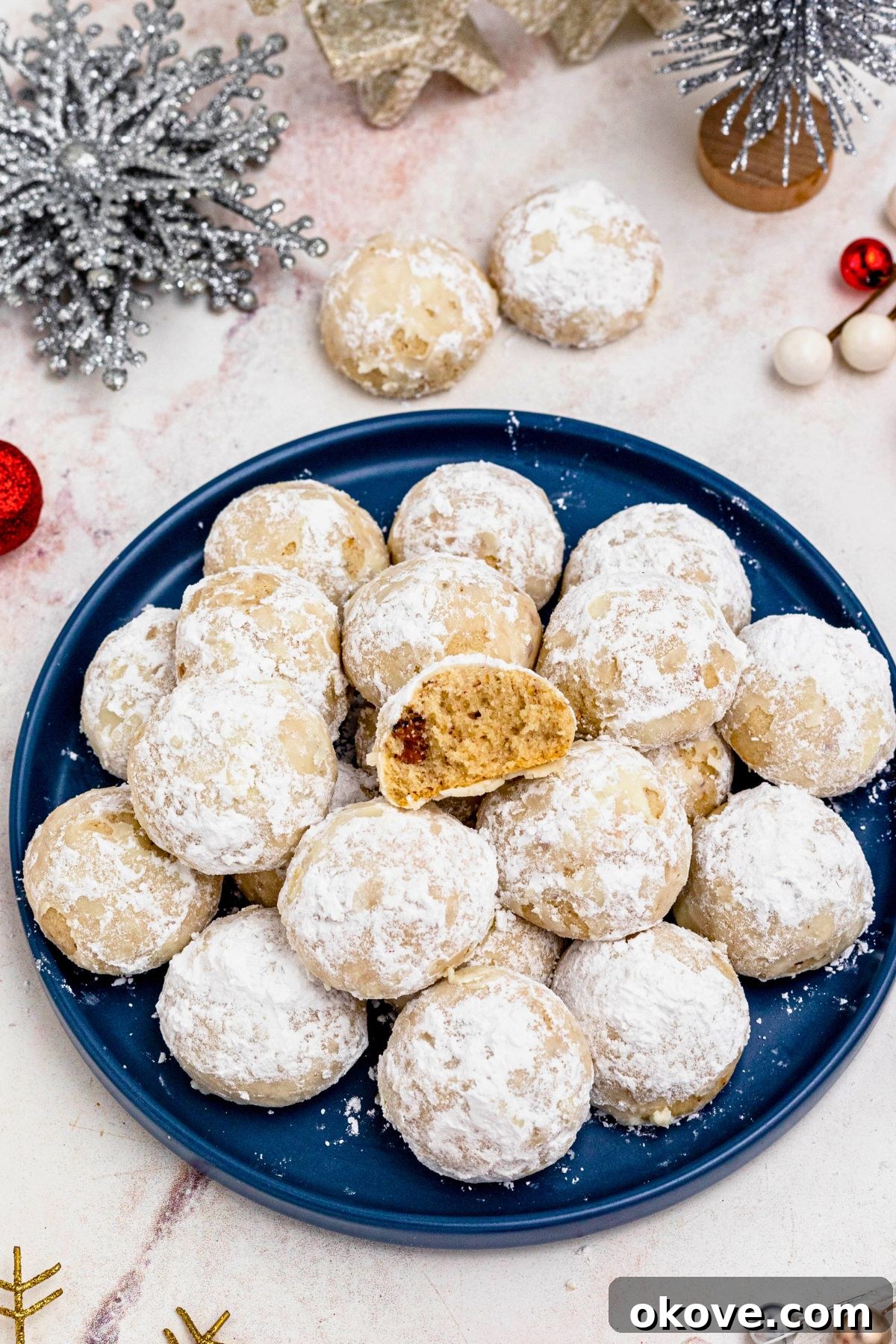 Powdered Cloud Kisses 10 Powder sugar coated Snowball cookies, stacked on a blue plate with holiday party favors on the table around the plate, with one cookie missing a bite.