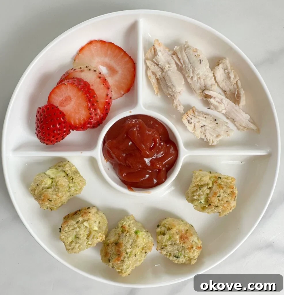 Crispy Parmesan Zucchini Bites 2 A white divided plate featuring golden brown zucchini parmesan tots, accompanied by crispy chicken pieces, a dollop of ketchup, and fresh sliced strawberries.