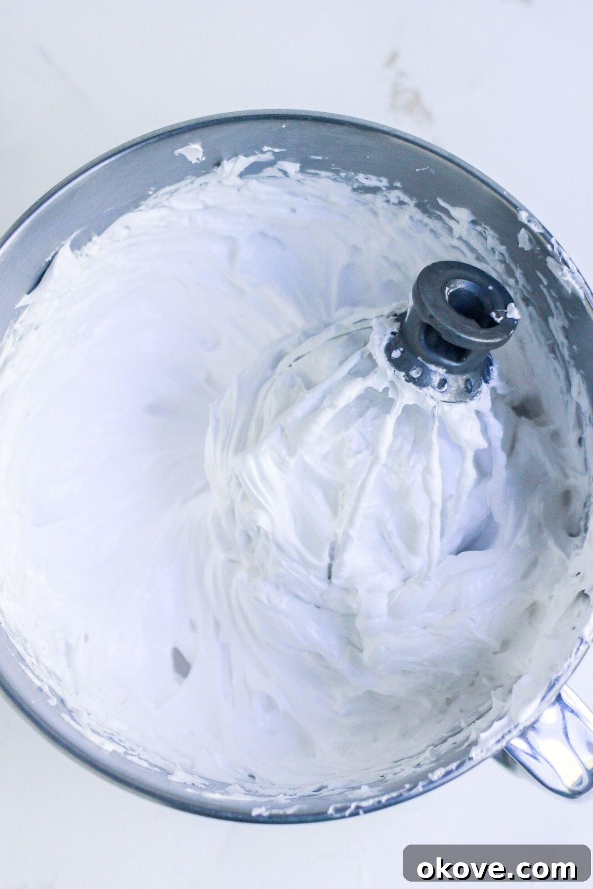 Egg whites, sugar, and other ingredients, mixed until thick and soft peaks form, in a silver mixing bowl. 