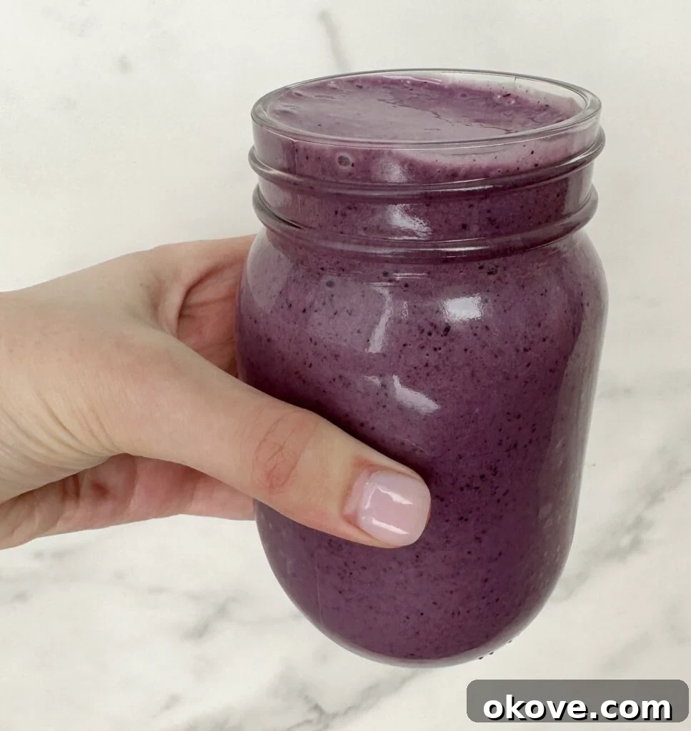 A hand holding a vibrant purple blueberry banana spinach smoothie in a clear glass jar, garnished with fresh blueberries.