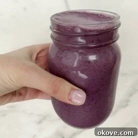 A hand holding a blueberry banana spinach smoothie in a clear jar