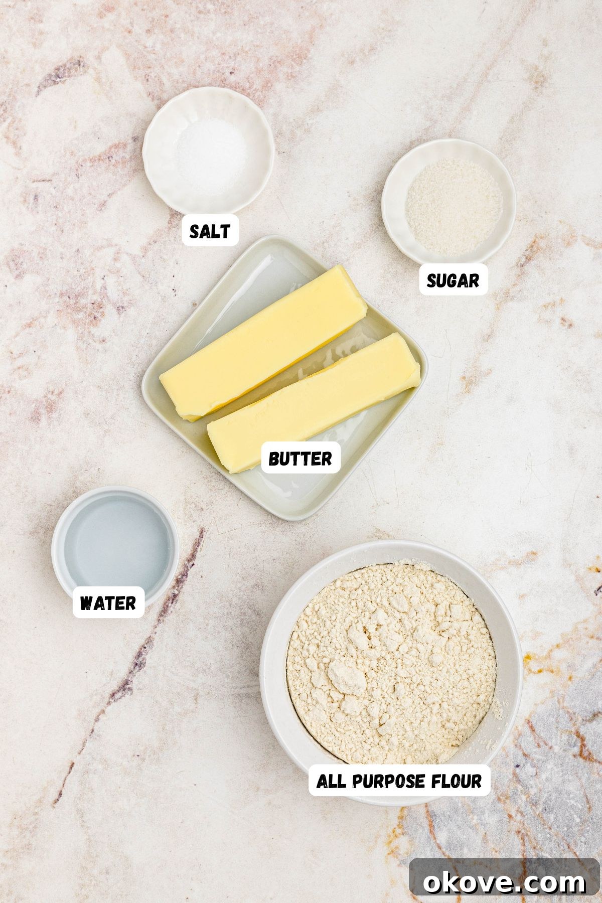 Flour, butter, water, salt, and sugar, measured into small bowls and labeled on a marble table.