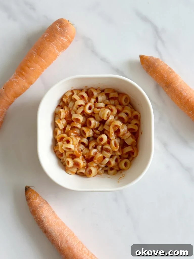 A white bowl filled with healthy homemade spaghetti-o's, garnished with fresh parsley, surrounded by whole, vibrant carrots and a wooden spoon.