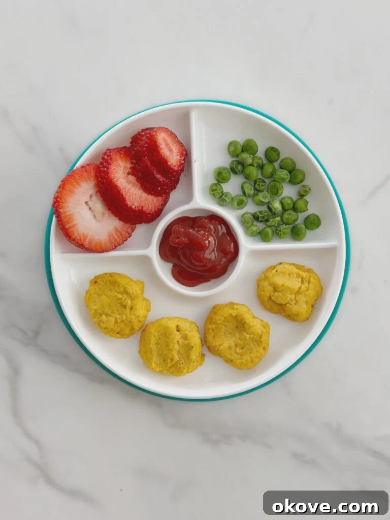 A divided plate with four carrot chicken nuggets, peas, sliced strawberries, and ketchup