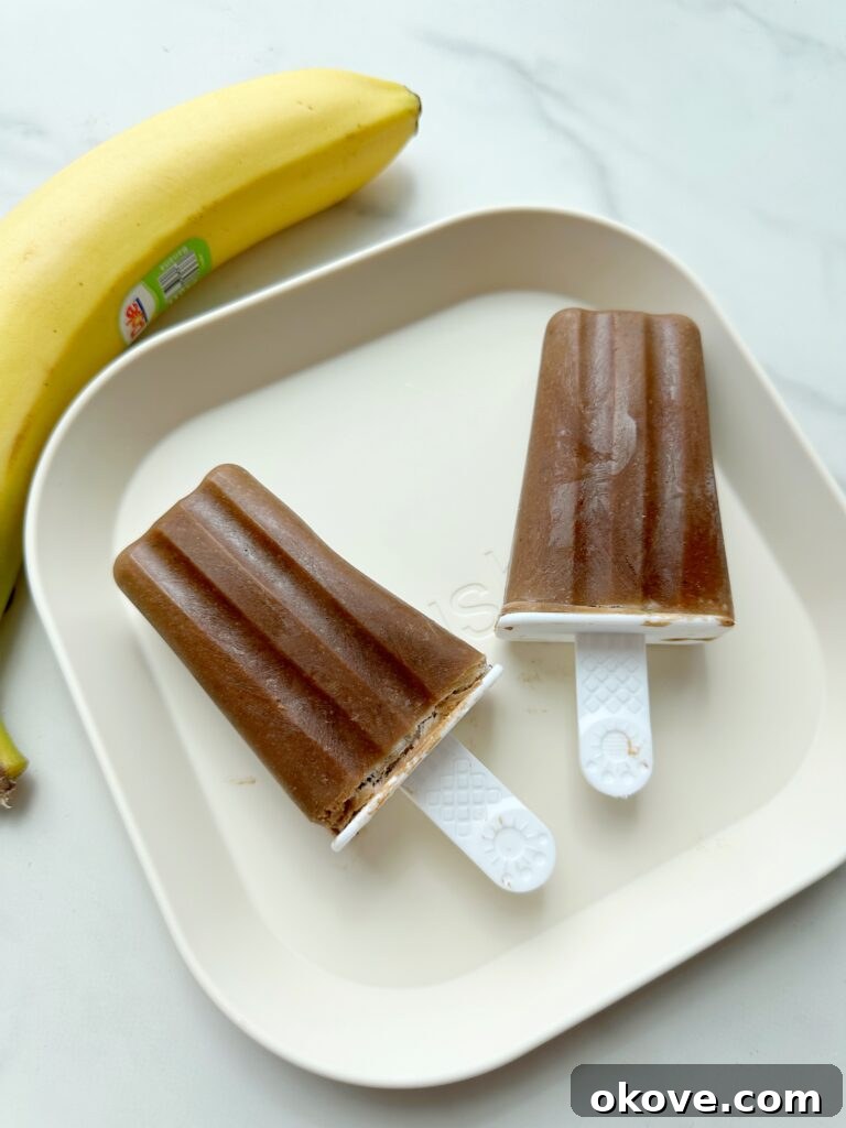 Guilt-Free Fudge Pops 2 Two brown fudgesicles on a white plate and a banana in the background, signaling a delicious and healthy treat for kids.