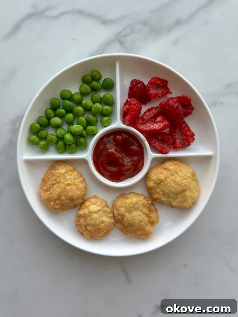 Four chicken nuggets on a white divided plate with raspberries and peas and ketchup