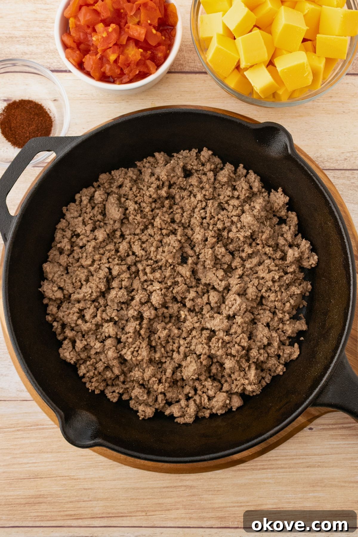 Rotel Fiesta Dip 4 Ground beef cooked in a black skillet with the grease drained and cheese cubes, chopped tomatoes, and seasonings on the table.