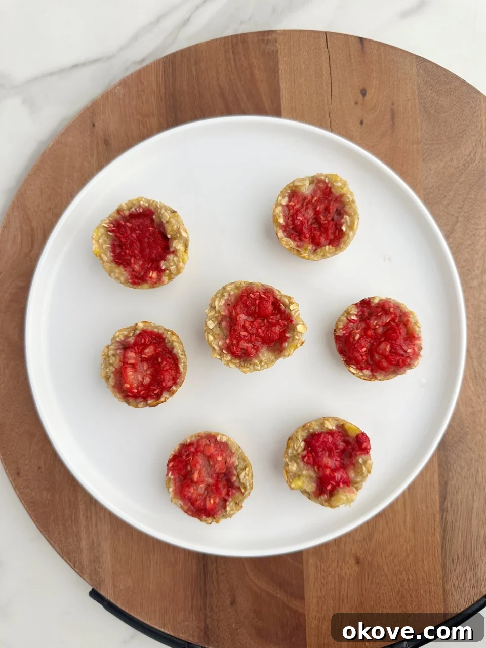Wholesome Raspberry Oat Bites 2 Seven raspberry oat bites on a white plate on a brown wood background, showcasing a healthy and easy toddler snack