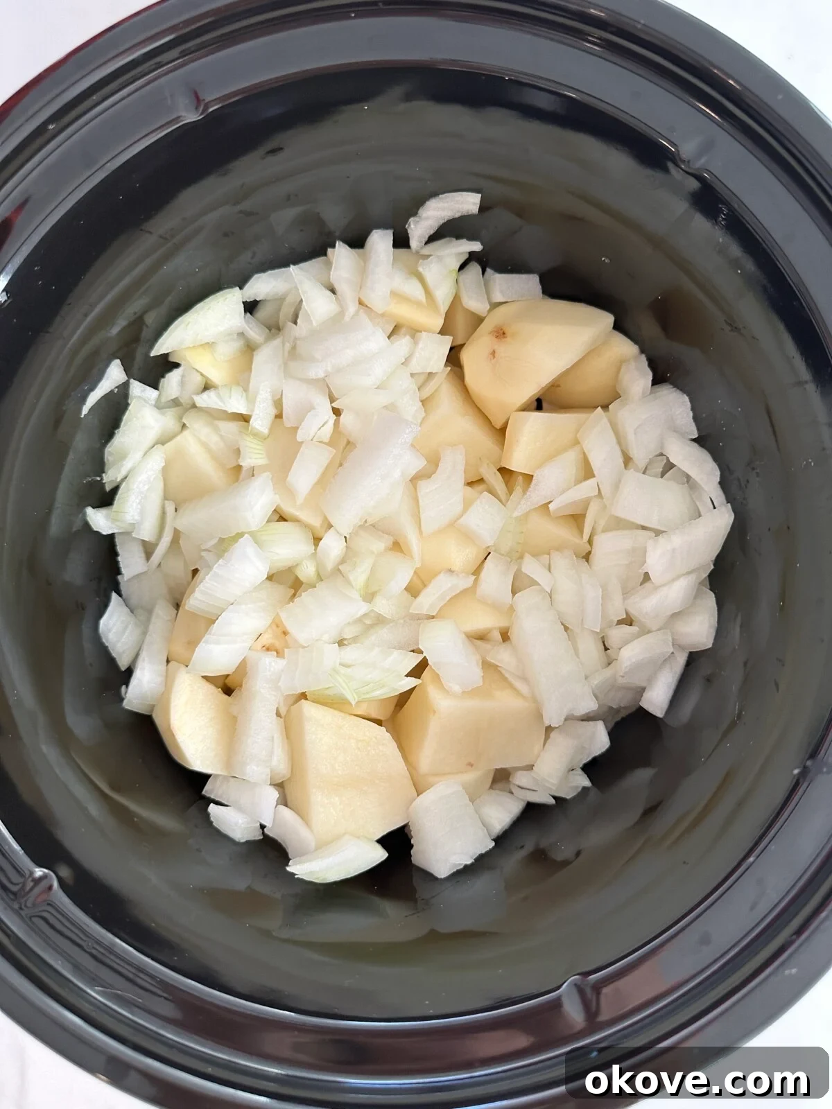 Peeled and diced potatoes and chopped onions submerged in chicken broth within a slow cooker, ready for cooking.