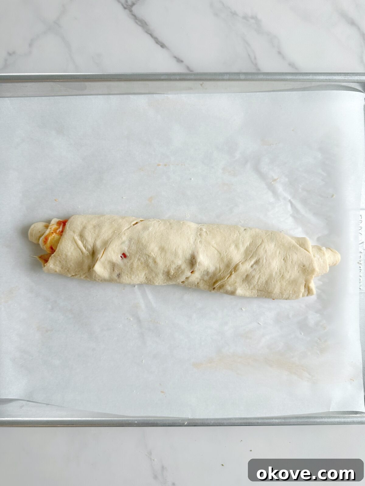 Savory Pizza Spirals 7 The crescent roll dough, fully topped with pizza ingredients, expertly rolled into a compact log shape, ready for slicing.