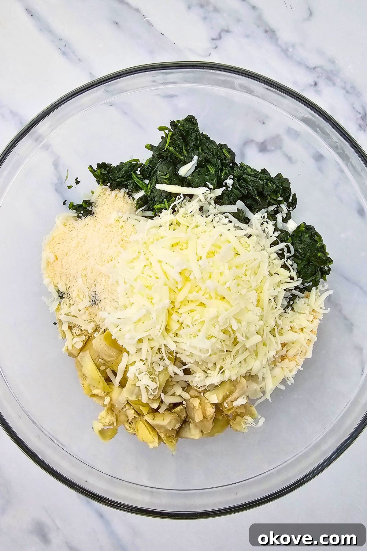 Creamy Spinach Artichoke Delight 4 Chopped spinach, artichoke hearts, and various cheeses in a large clear glass mixing bowl before being combined.