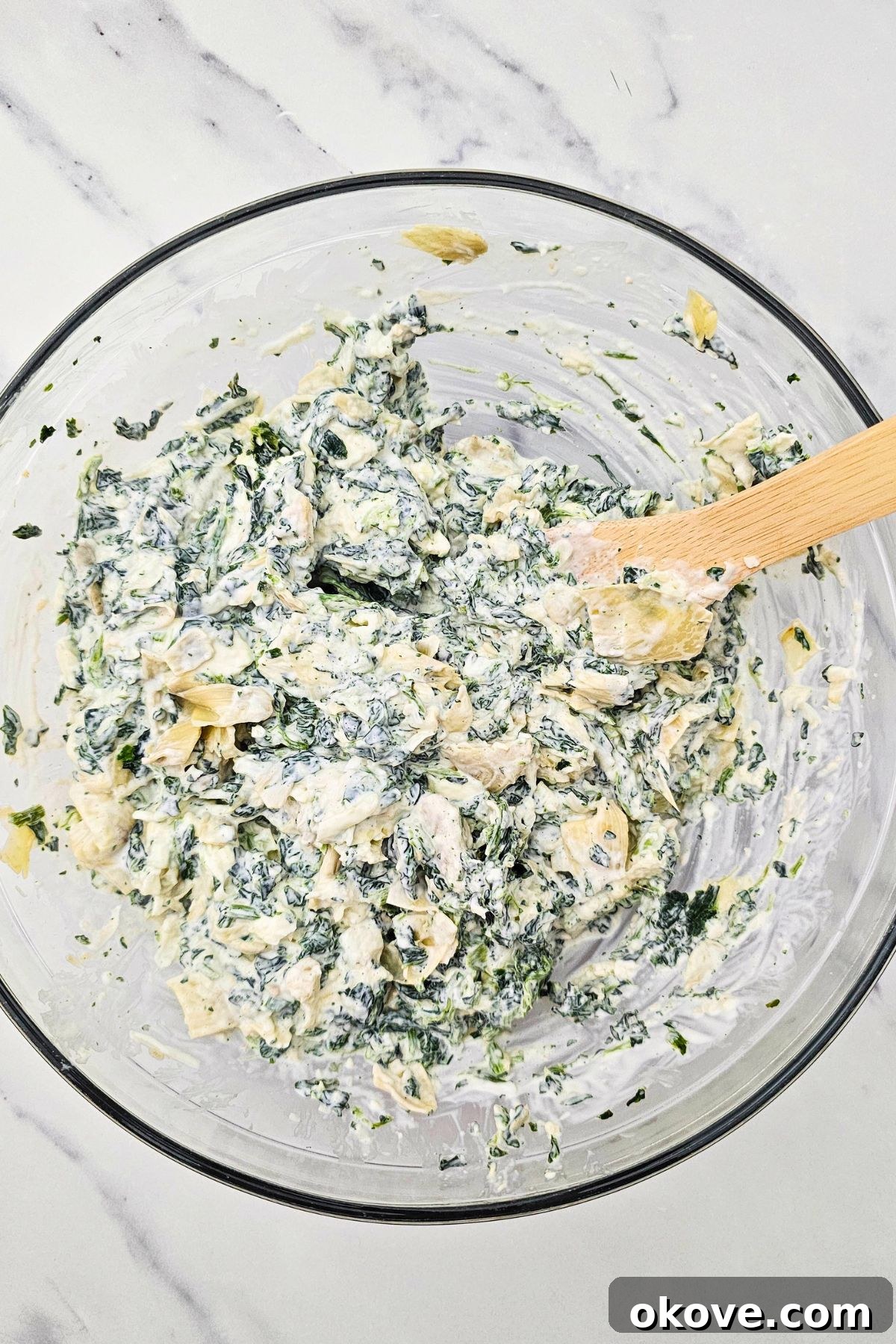 Creamy Spinach Artichoke Delight 5 The spinach, artichoke, and cheese mixture being mixed together with a wooden spoon in a large clear glass bowl until well combined.