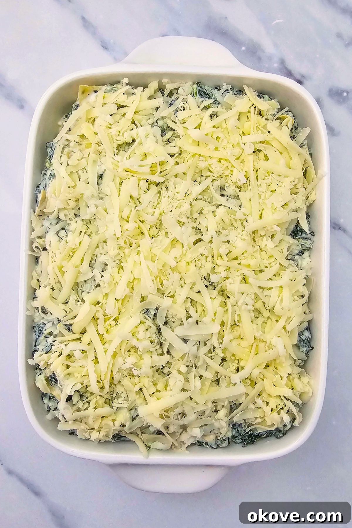 Creamy Spinach Artichoke Delight 6 The uncooked spinach and artichoke mixture spread evenly into a rectangle baking dish, topped with the remaining shredded cheese, on a marble counter.
