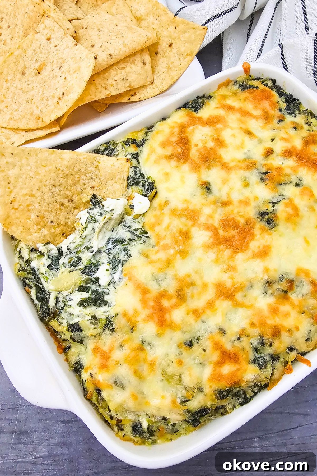 Creamy Spinach Artichoke Delight 7 Warm, creamy spinach artichoke dip in a square baking dish, with a tortilla chip scooping out a portion, ready to serve.