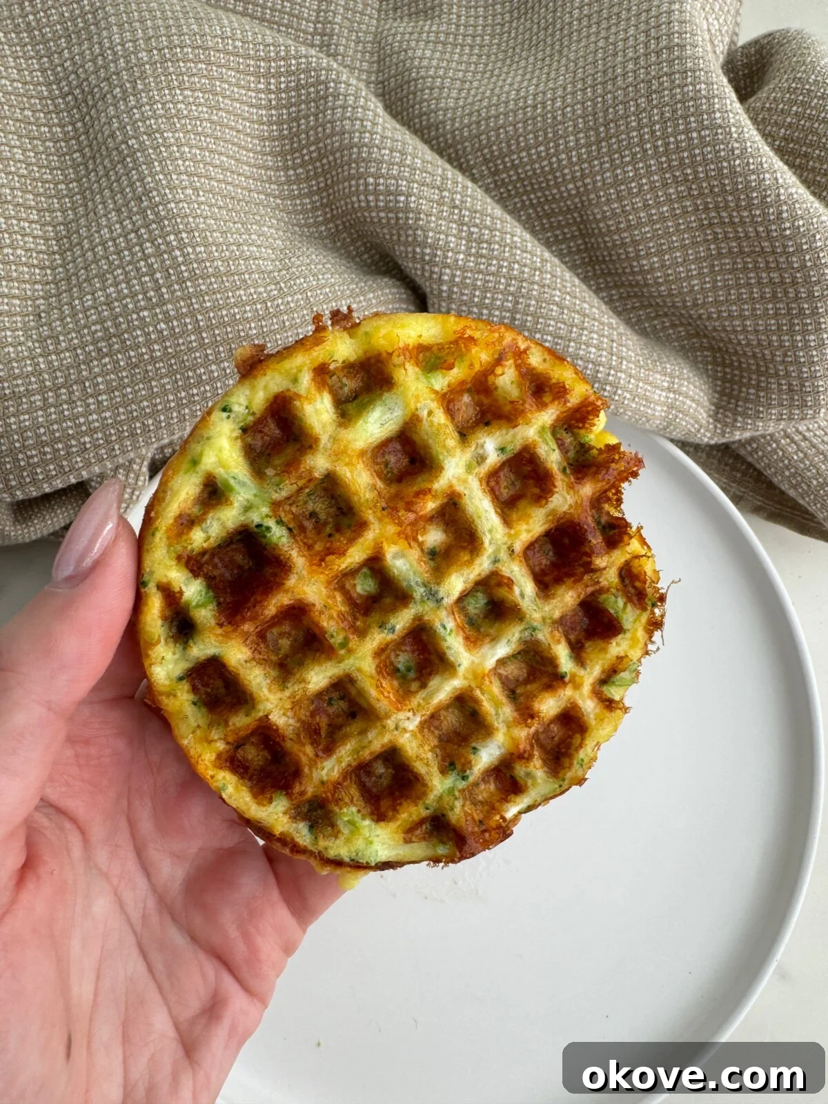 A hand holding up a broccoli cheese waffle, showing its golden-brown texture and green flecks of broccoli.