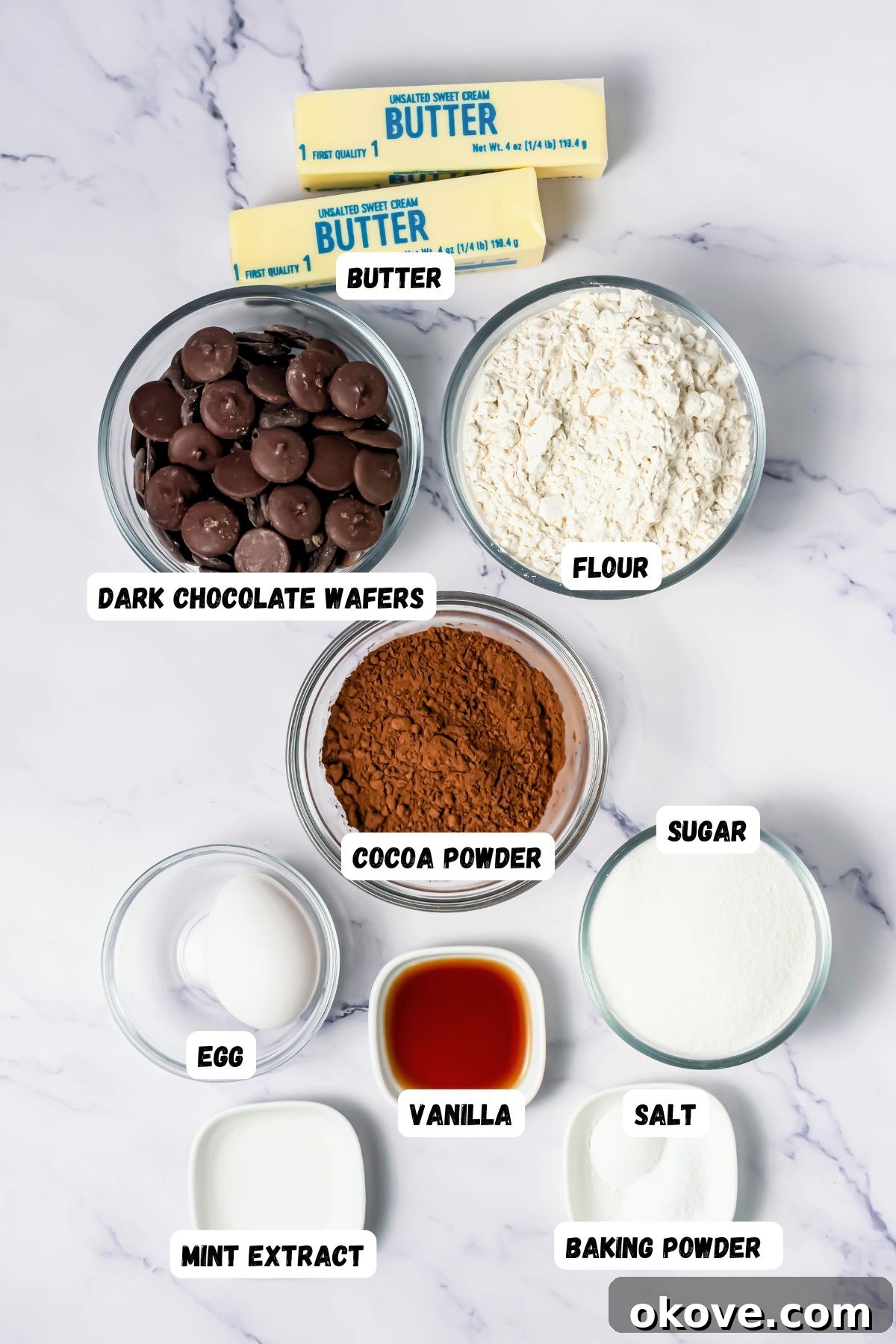 Ingredients like flour, sugar, butter, and more, measured into small bowls on a white marble table and labeled. 