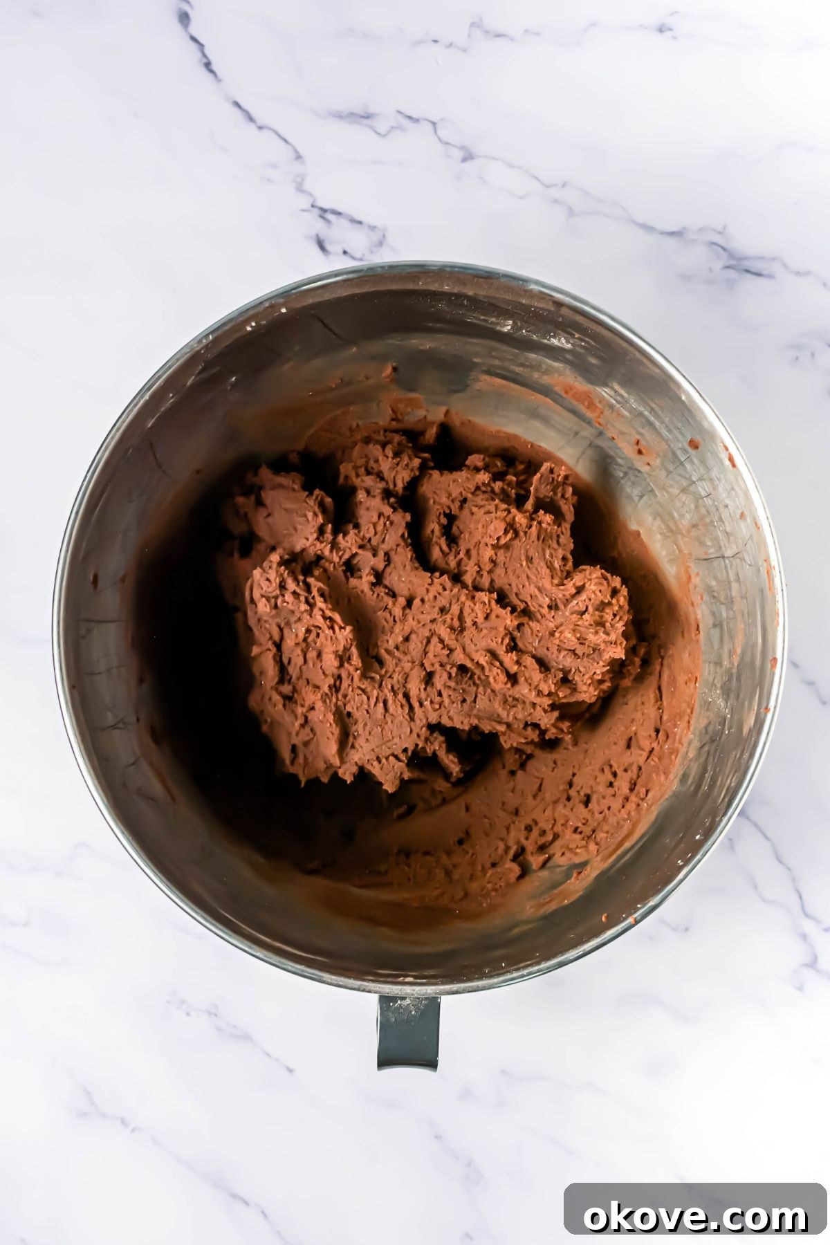 Chocolate cookie dough, formed in a silver mixing bowl, on a white marble table.