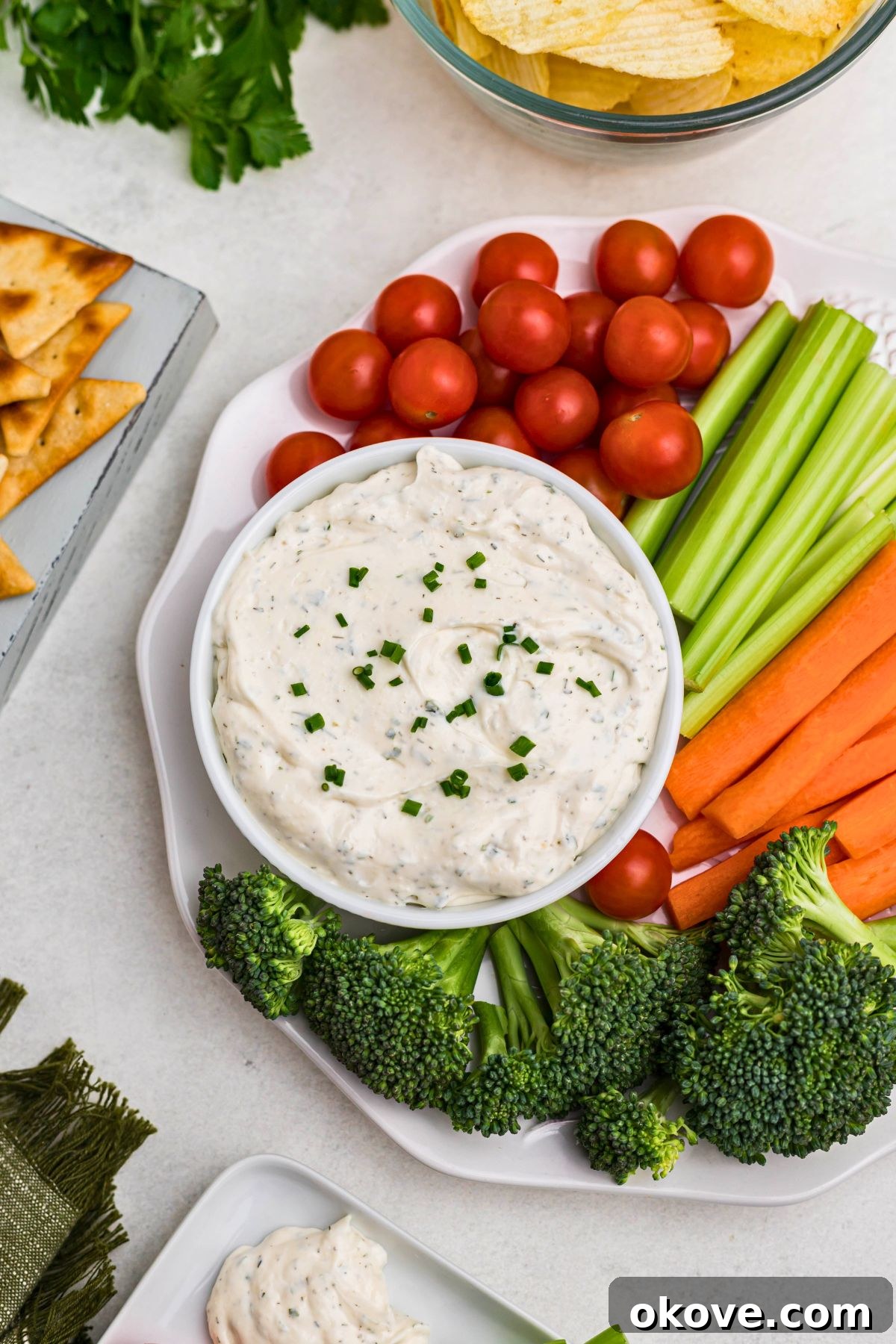 Creamy Ranch Dip 2 Small white bowl filled with creamy ranch dip, elegantly surrounded by an assortment of fresh, colorful vegetables like carrots, celery, broccoli, and bell peppers on a white plate. A separate plate with crispy potato chips and pita chips completes the inviting display.
