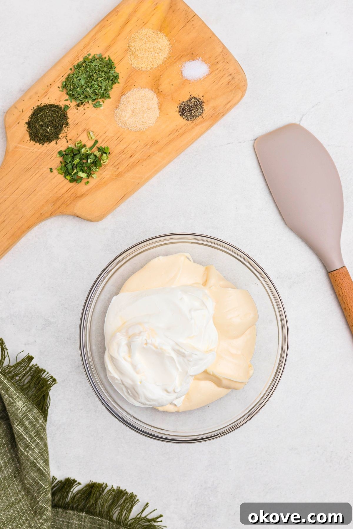 Creamy Ranch Dip 4 A clear glass bowl on a cutting board holds creamy mayonnaise and sour cream, with various dried herbs and spices neatly arranged around it, poised for mixing.