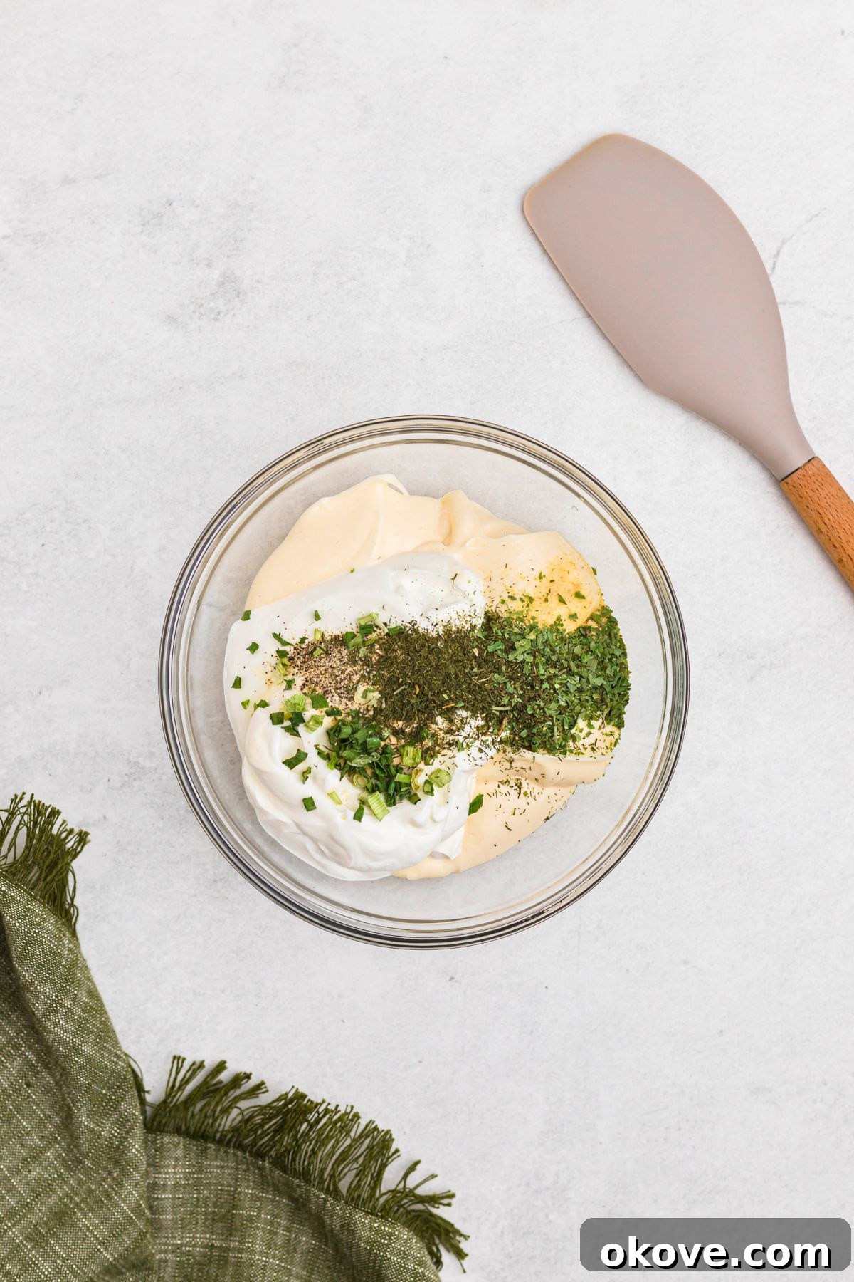 Creamy Ranch Dip 5 The dried herbs and seasonings, including dill, parsley, and chives, are sprinkled over the creamy base of mayonnaise and sour cream in a clear glass bowl, ready for the next mixing stage.
