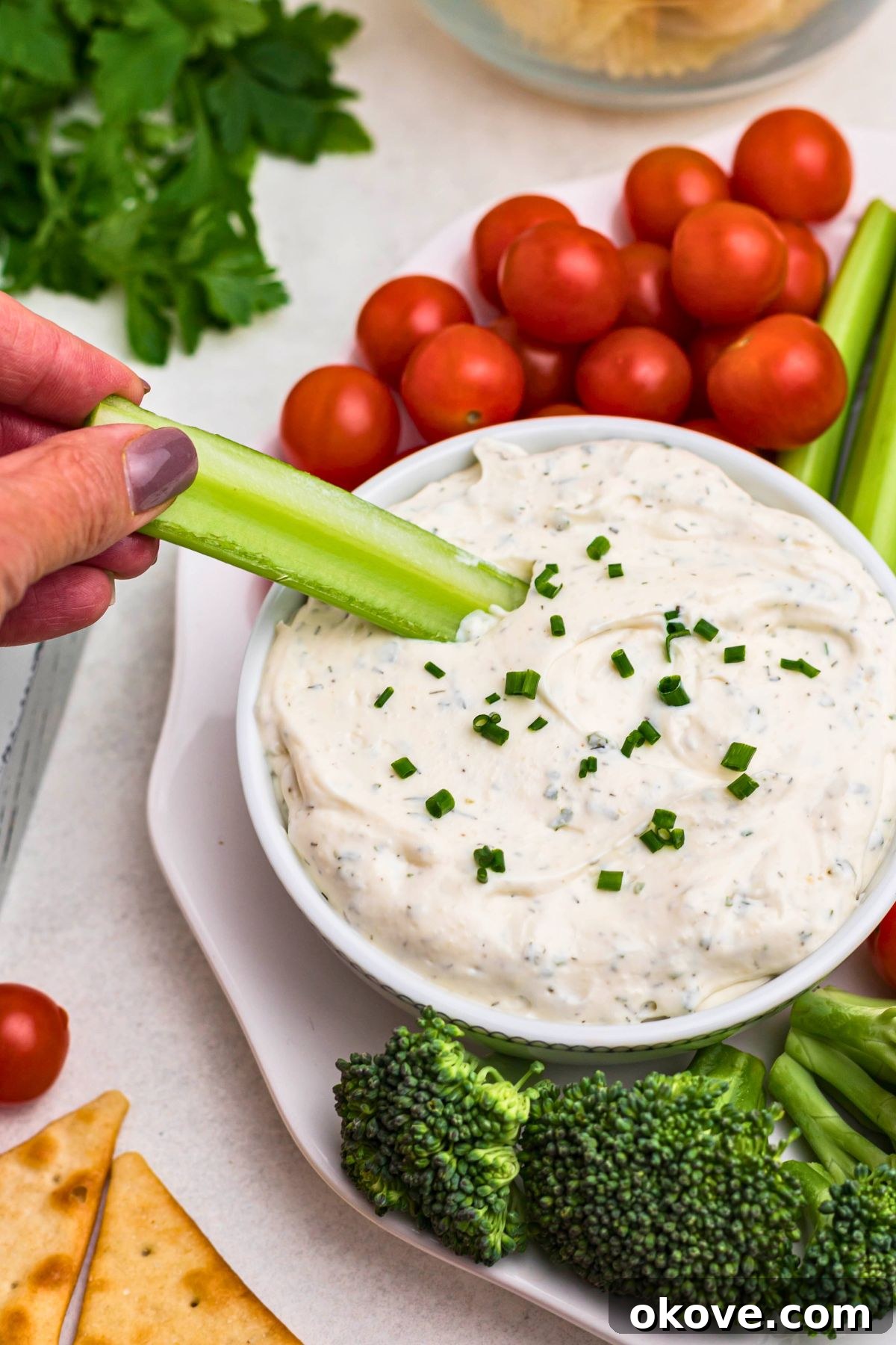 Creamy Ranch Dip 7 A small white bowl filled with creamy ranch dip is centered on a white plate, surrounded by an array of fresh vegetables and crispy chips. A hand is reaching into the dip with a crisp celery stick, highlighting the perfect consistency for dipping.