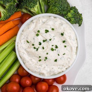 Small white bowl filled with dip, surrounded by fresh vegetables on a white plate, on table.