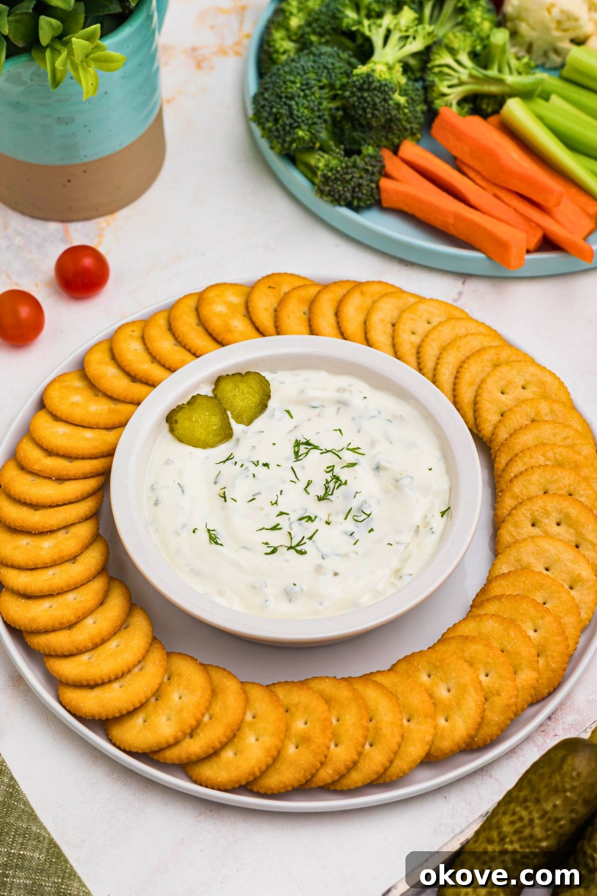 Small white bowl filled with creamy white dip, topped with slices of pickles, surrounded by round crackers, and a plate of fresh vegetables. 