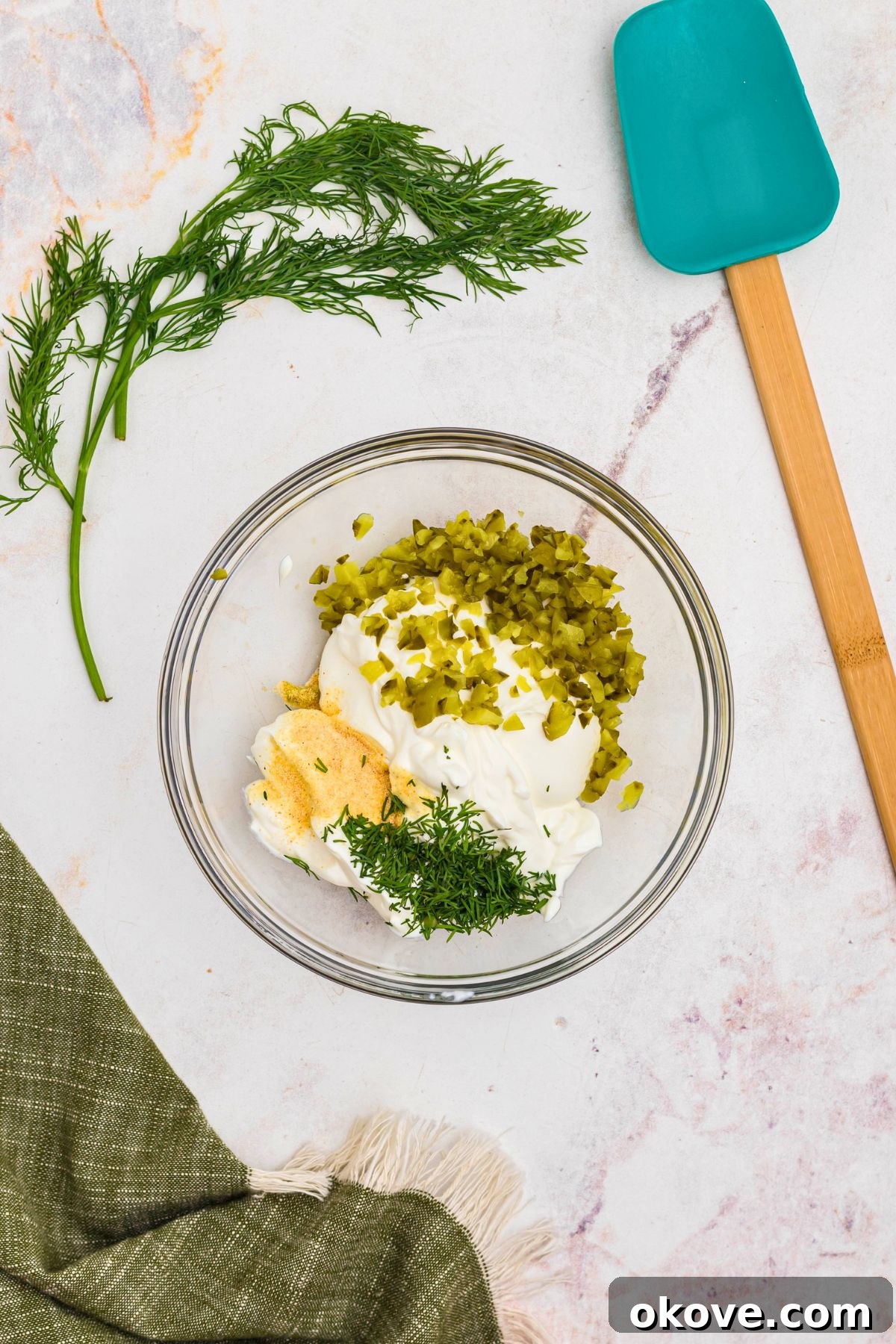 Sour cream, chopped pickles, and other ingredients, in a clear glass bowl, before being mixed together.