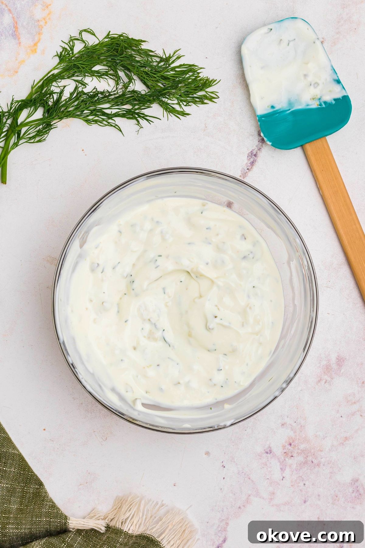Creamy dip mixed together in a clear glass bowl with a spatula and fresh dill laying on the table next to the bowl. 