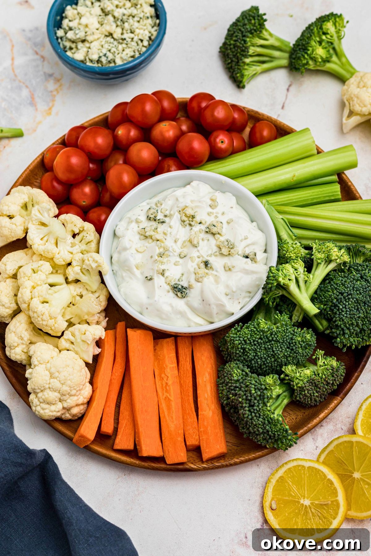 Creamy Blue Cheese Dip 2 A small round white bowl filled with creamy blue cheese dip, generously topped with crumbled cheese, and surrounded by a vibrant assortment of freshly chopped vegetables like celery, carrots, and cucumber. A piece of celery is being dipped into the center, highlighting the dip's rich texture.