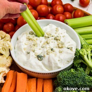 A small round white bowl filled with creamy blue cheese dip, topped with crumbled blue cheese, and surrounded by an assortment of fresh vegetables, including a celery stick being dipped into the center.