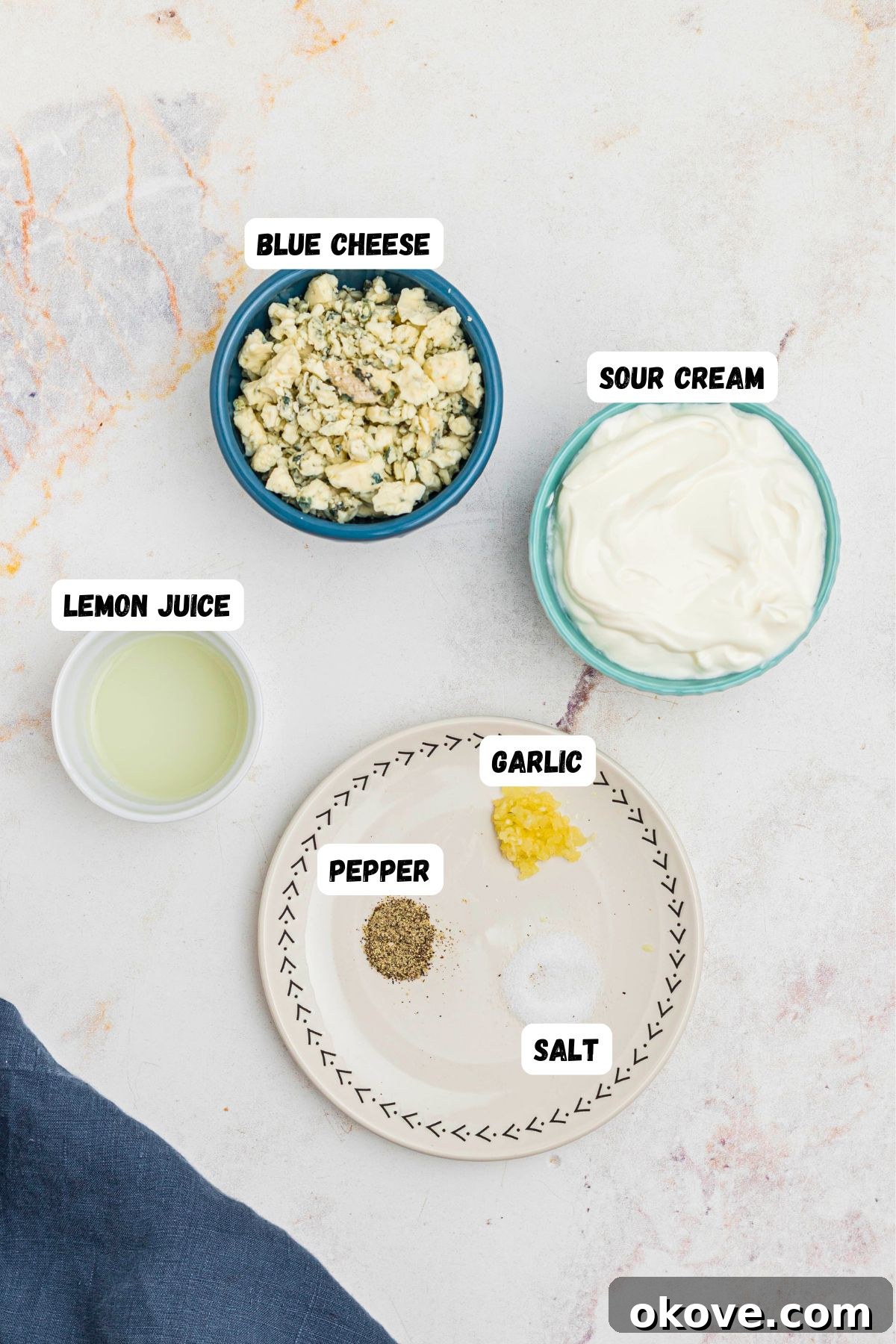 Creamy Blue Cheese Dip 3 Small bowls on a marble table containing the individual ingredients for blue cheese dip: sour cream, a wedge of blue cheese, fresh lemon juice, and minced garlic, all clearly labeled.