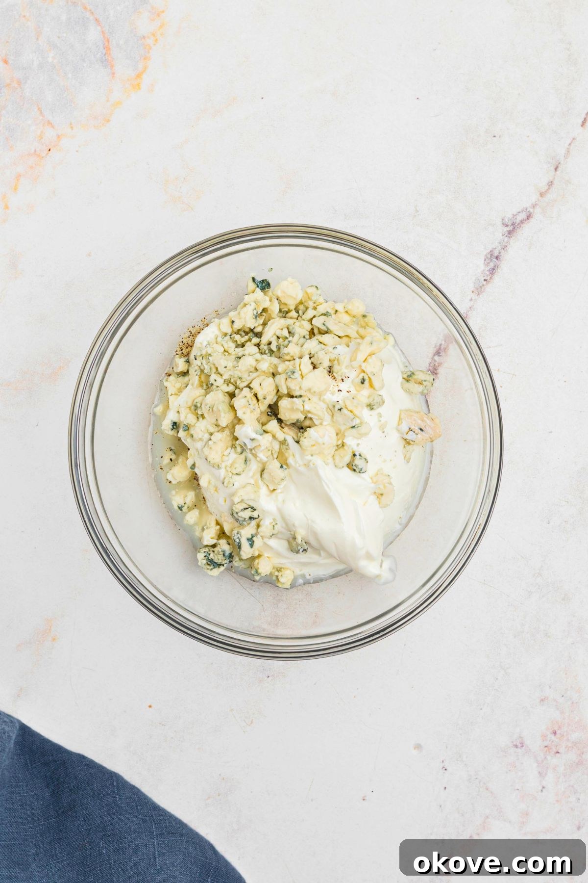 Creamy Blue Cheese Dip 4 A clear glass mixing bowl containing sour cream, crumbled blue cheese, lemon juice, and minced garlic, positioned on a kitchen counter before being combined.