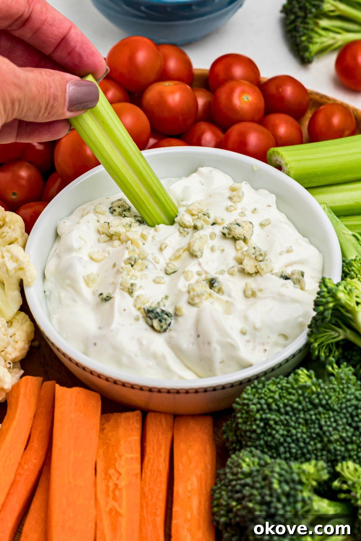 Creamy Blue Cheese Dip 6 A beautifully presented white round bowl brimming with creamy blue cheese dip, artfully mixed with cheese crumbles and seasonings. It is surrounded by a colorful array of fresh vegetables, and a crisp celery stick is being dipped into the rich center, inviting a taste.