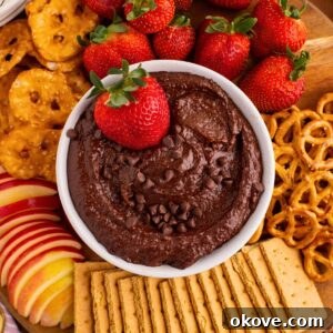 A pristine white bowl filled with rich, decadent chocolate hummus, with a golden serving spoon poised in the center. The bowl is artfully surrounded by an array of delectable dipping items, including fresh, juicy strawberries and salty pretzels.