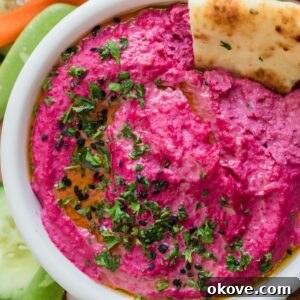 A bowl of vibrant pink beet hummus with a cracker delicately placed within it, garnished with a sprinkle of fresh parsley flakes for extra appeal.