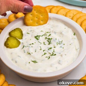 A small white bowl filled with creamy white dip, generously topped with thin slices of dill pickles. It is surrounded by a bounty of round crackers and a diverse plate of fresh vegetables, ready for dipping.