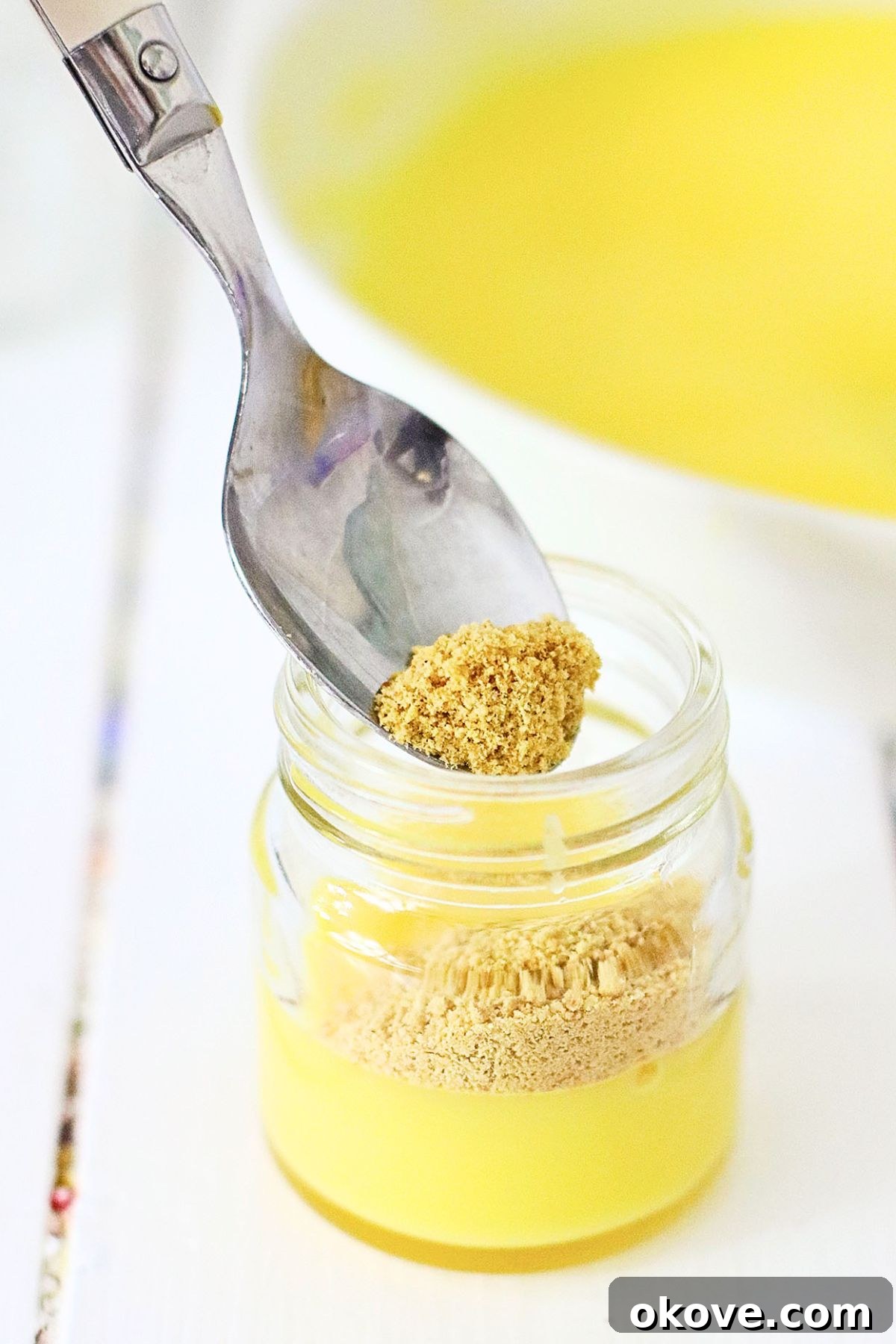 Sweet Banana Pudding Shooters 5 A person carefully spooning a layer of crushed cookies into a mini shot glass, creating a distinct layer over the bright yellow banana pudding mixture.